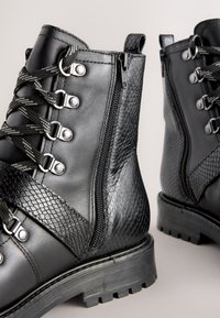 Musta nahkaiset nilkkurit, joissa on teksturoitu sivupaneeli, vetoketjusuljenta, metalliset nyöriholkit ja pito pohjassa.