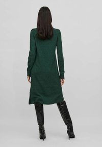 Vestito verde a maglia che arriva fino al polpaccio con spacco laterale. Maniche lunghe, texture morbida e vestibilità rilassata. Abbinato a stivali neri al ginocchio.