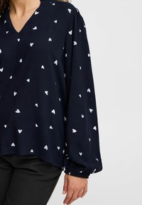 Blouse bleu marine avec motif de cœurs blancs, décolleté en V, manches longues bouffantes et poignets élastiques. Tissu doux et léger avec une coupe décontractée.