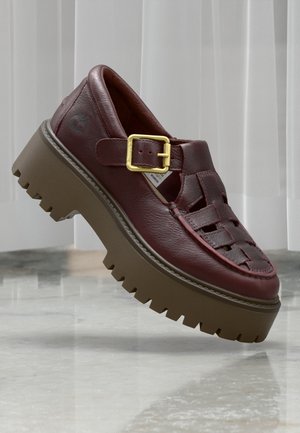 Kraftig brun læder sandal med guldlås og tyk riflet sål, sat op mod lyse lodrette persienner og marmorgulv.
