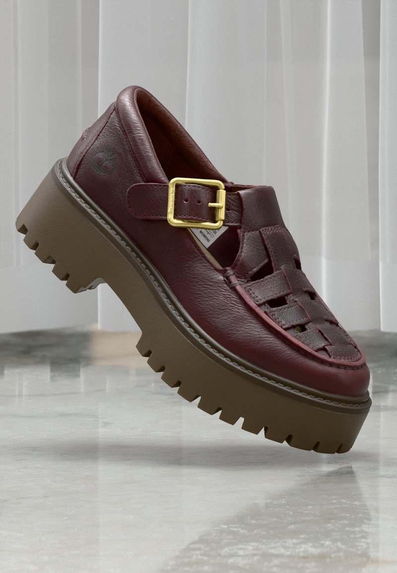 Sandale en cuir marron épais avec boucle dorée et semelle épaisse à rainures, placée devant des stores verticaux clairs et un sol en marbre.