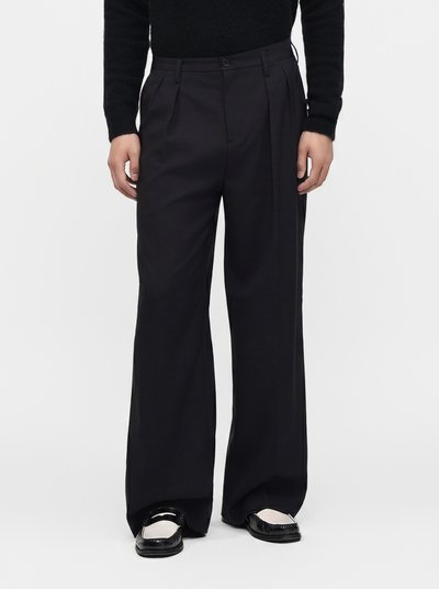 Person wearing loose-fitting black trousers and black and white loafers, standing against a plain light background.