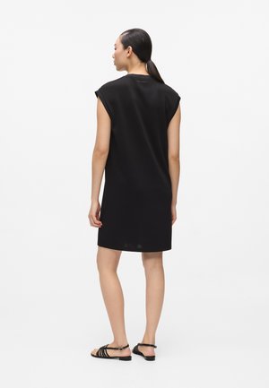 Frau steht mit dem Rücken zur Kamera, trägt ein ärmelloses, knielanges schwarzes Kleid und schwarze, flache Sandalen mit Riemchen vor einem schlichten weißen Hintergrund.