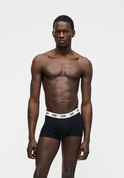 Μυώδης άνδρας που στέκεται μπροστά σε λευκό φόντο, φορώντας μαύρα εσώρουχα τύπου μποξέρ με λευκή μέση και το λογότυπο "GCDS", κοιτάζοντας μπροστά.
