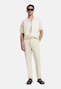 Hellbeige Kurzarmhemd mit gemusterter Textur, kombiniert mit passenden, schmal zulaufenden Hosen und schwarzen Sandalen, ergänzt durch eine Sonnenbrille.
