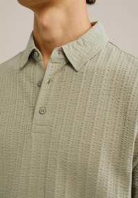 Camisa de color verde claro de manga corta hecha de tela texturizada, con cuello, tres botones y un detalle de bolsillo en el pecho.