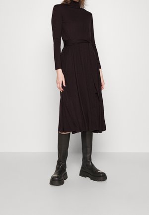 Kobieta ubrana w czarną midi sukienkę z długim rękawem i paskiem w talii oraz czarne masywne kozaki za kolano na tle jednolitego tła.