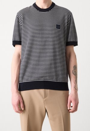 Mann trägt ein schwarz-weiß horizontal gestreiftes kurzärmeliges Hemd mit beigen Hosen, zeigt ein Tattoo am rechten Unterarm.