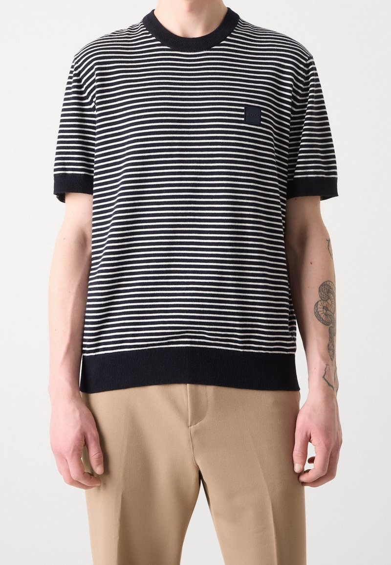 Homme portant un t-shirt à manches courtes rayé horizontal noir et blanc avec un pantalon beige, montrant un tatouage sur l'avant-bras droit.