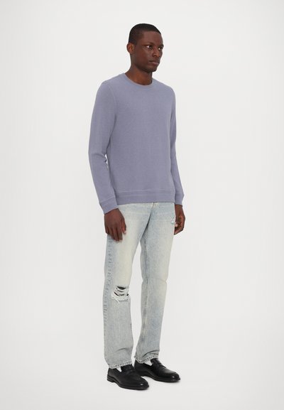Pull violet à manches longues avec un col rond, porté avec un jean bleu clair déchiré et des chaussures noires, présentant une coupe décontractée et une texture douce.