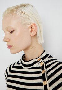 Črtast pleten pulover z visokim ovratnikom, črno-krem barvami in zlatimi dekorativnimi gumbi ob rami.