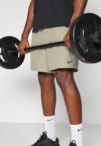 Zwarte Nike t-shirt, licht olijfgroene shorts, houdt een zwarte halterstang met gewichten vast. Zwarte sneakers en witte geribbelde sokken zichtbaar.
