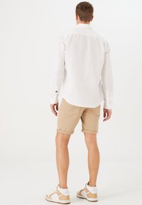 Weißes, langärmliges Leinenhemd kombiniert mit beigen, hochgerollten Shorts und hellbraunen Sneakern mit weißen Akzenten. Einfaches, lässiges Outfit.
