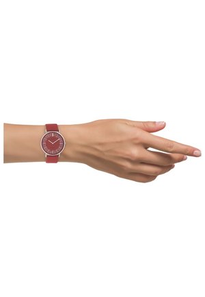 Main féminine portant une montre-bracelet rouge avec un cadran texturé dans un boîtier en argent, présentée sur un fond blanc.
