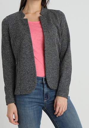 Vrouw draagt een nauwsluitend, gestructureerd grijs colbert over een roze shirt en blauwe jeans, staand met één hand rustend nabij haar heup.