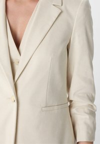 Veste couleur crème, en tissu texturé, avec un col à revers cranté, deux boutons à l'avant et une poche poitrine unique.