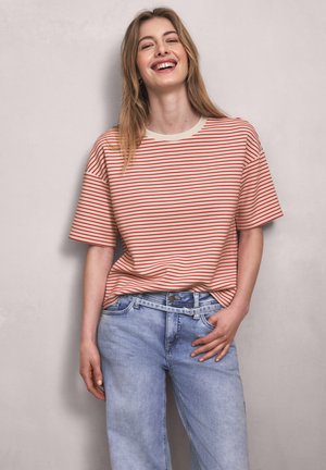 Femme souriante aux cheveux longs portant un T-shirt rouge et blanc à rayures et un jean bleu clair, debout devant un mur uni.