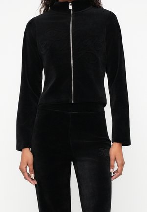 Frau trägt schwarze Velours-Zipjacke und passende Hose mit dezentem, besticktem Logo auf Brust und Oberschenkel.