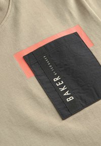 Beige bomulds t-shirt med en fremtrædende sort stofpatch med et hvidt logo og en rød rektangulær accent over lommen.