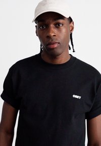 Zwarte t-shirt met opgerolde mouwen toont een wit "OBEY" logo op de voorkant. Het model draagt een lichtbeige pet. Effen achtergrond.