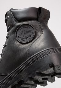 Botte Palladium en cuir noir avec patch logo rond, col rembourré et semelle crantée épaisse, vue de côté et de dessous.