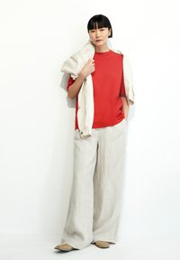 Rode top met korte mouwen, beige linnen wijde broekspijpen en beige instapschoenen. Lichtgekleurde jas over de schouder gedrapeerd. Neutrale achtergrond.