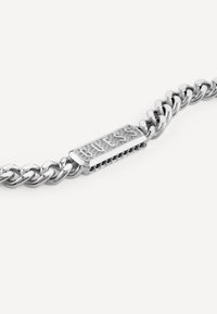 Srebrna bransoletka z łańcuchem, z prostokątnym zapięciem z wygrawerowaną nazwą marki, gładkimi ogniwami i polerowanym wykończeniem. Podkreśla błyszczącą metaliczną teksturę.