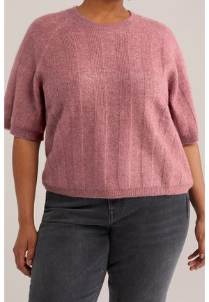 Osoba ubrana w różowy, prążkowany sweter z krótkim rękawem oraz ciemnoszare dżinsy, stojąca na tle jednolitego tła.