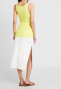 Débardeur jaune ajusté avec un décolleté rond, associé à une jupe midi blanche avec une fente sur le côté et des détails de couture visibles. Sandales à glissière marron.