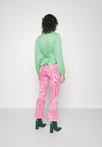 Zelená texturovaná blůza s nabíranými rukávy, v kombinaci s růžovými vzorovanými kalhotami a zelenými lakovanými kotníkovými botami, viděná zezadu.
