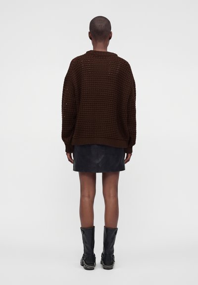 Personne debout dos à la caméra portant un pull en grosse maille marron, une jupe en cuir noire et des bottes noires mi-mollet avec des détails en chaîne.