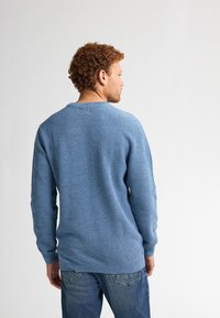 Pulover tricotat texturat albastru cu mâneci lungi și guler rotund, asociat cu blugi din denim. Țesătura are un model subtil și o croială relaxată.