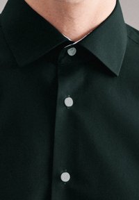 Mężczyzna w czarnej koszuli formalnej zapinanej na guziki z białymi guzikami, okazywany jest dekolt i górna część klatki piersiowej.