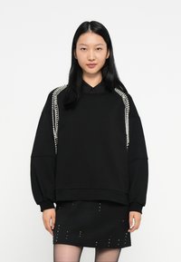 Žena s dlhými čiernymi vlasmi, oblečená v čiernom oversized svetri s perlovou výzdobou na ramenách a čiernej sukni s malými študmi.
