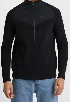 Czarna kurtka Nike na zamek z siateczkowymi panelami na ramionach, noszona przez osobę w niebieskich dżinsach, stojącą na jednolitym jasnym tle.