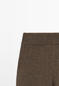 Close-up van bruin gebreide stof met een brede geribbelde tailleband, met zichtbare textuur en breipatroon tegen een witte achtergrond.