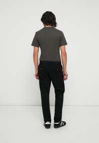 Camicia grigia a maniche corte abbinata a pantaloni neri a gamba dritta e sneaker scure, vista da dietro su uno sfondo neutro.