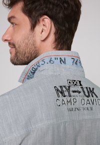 Camisa de botones a rayas en azul claro con textura de denim. Presenta texto bordado en el cuello y la parte posterior, incluyendo "NY UK CAMP DAVID."