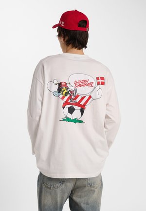 Person trägt weißes Langarmshirt mit einer gezeichneten Biene in dänischer Fußballkleidung, tritt Ball, rote Kappe und dänische Flagge auf Shirt-Rücken.