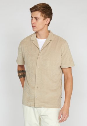 Kurzärmeliges Hemd in Beige, aus weichem Frottee-Stoff, mit Knopfleiste und Camp-Kragen, kombiniert mit hellen Hosen.