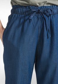 Tmavě modré kalhoty s volnějším střihem, elastickým pasem a stahovací šňůrkou. Materiál je hladký s jemným leskem. Viditelné boční kapsy.