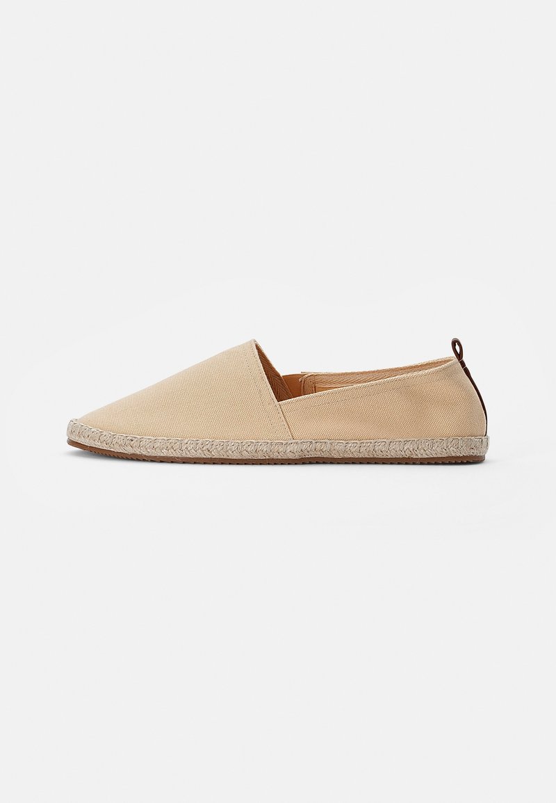 Espadrille beige à enfiler avec dessus en toile, bordure de semelle en jute tressée et languette marron au talon, présentée de profil sur fond blanc.