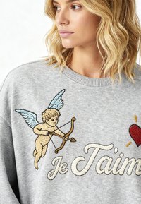 Femme blonde portant un pull gris avec un chérubin brodé tenant un arc et les mots "Je T’aime" accompagnés d'un cœur rouge.