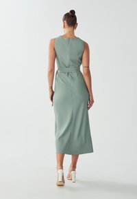 Mint green sleeveless midi dress, smooth texture, fitted waist with tie, zipper closure, and subtle seam detailing. Heels visible.