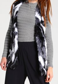 Czarny i biały kamizelka z sztucznego futra o długim, prostym kroju. Kamizelka ma miękką teksturę i wyraźny wzór pasków, noszona na paskowanym topie.