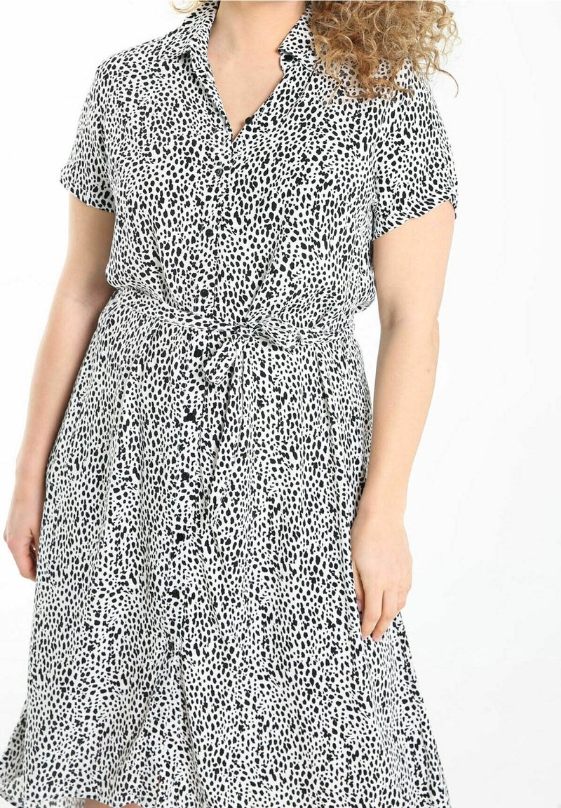 Femme portant une robe à manches courtes, à pois noir et blanc, boutonnée avec une ceinture nouée à la taille, sur fond blanc.