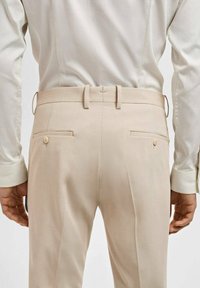Bež hlače s krojem po meri, ki imajo dve zadnji žepi z gumbi in ozkimi pasovi. Gladka tekstura in minimalističen dizajn.