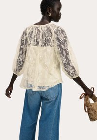 Elfenbeinfarbene Spitzenbluse mit Puffärmeln und rundem Ausschnitt, die mit Blumenmustern verziert ist. Kombiniert mit hellblauen weit geschnittenen Jeans und einer geflochtenen Tasche.