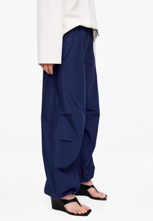 Personne portant un pantalon cargo ample bleu marine, un haut blanc à manches longues et des sandales compensées noires à orteils ouverts, debout de côté.