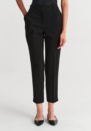 Femme portant un pantalon noir ajusté longueur cheville avec des plis à l’avant et des ballerines pointues noires, debout avec une main dans la poche.